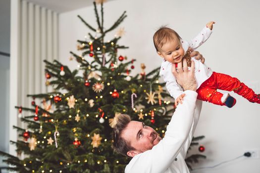 Father and daughter, baby girl having fun on Christmas, decorating the Christmass tree in festive outfits. Smiling baby girl playing with her dad.