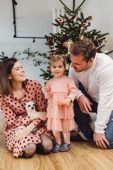 Beautiful young caucasian family Christmas portrait. Mom, dad and a little girl. Mom and daughter wearing pink dresses, dad wearing a white sweater. A little dog, chihuahua joining them on the photos. Family portrait in front of the Christmas tree.