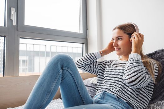 Blonde young caucasian woman lying on her dorm room bed next to a window putting on headphones, listening to music on a cold cloudy autumn day.