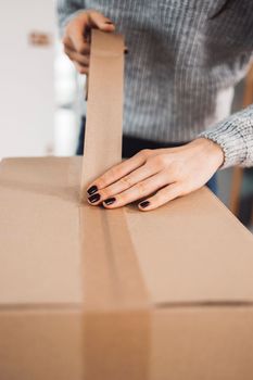 Young caucasian woman wrapping a gift, getting it ready to send in the post. Woman packaging a surprise gift for a family member, packaging it into a brown cardboard box.