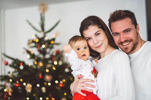 Happy young cheerful caucasian family of three mom dad and baby girl having fun decorating the Christmas tree. Family Christmas portrait.