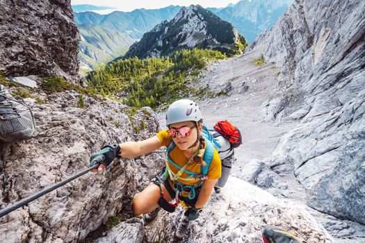 Caucasian woman mountaineer climbing up the mountain on via ferrata trail, high up in the mountain peaks. Woman wearing a protective head gear and uses a safety harness while she climbs.