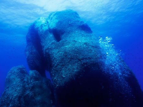 Underwater stone, Pietra di Saragossa, Sardinia. The large rock reaches from the seabed to the surface illuminated by the sun. Small fish swim around, some bubbles go up on one side