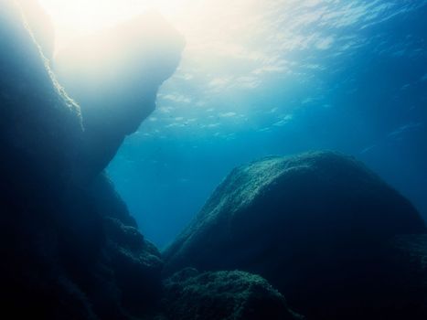 The dark rocks of the seabed are backlit by sunlight, this is reflected in the golden surface of the crystal blue water
