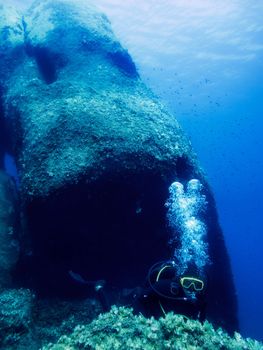 A person dives near the seabed next to a huge rock known as Pietra di Saragossa. The bubbles of the diver's breathing rise to the surface illuminated by sunlight. Small fish swim next to the stone