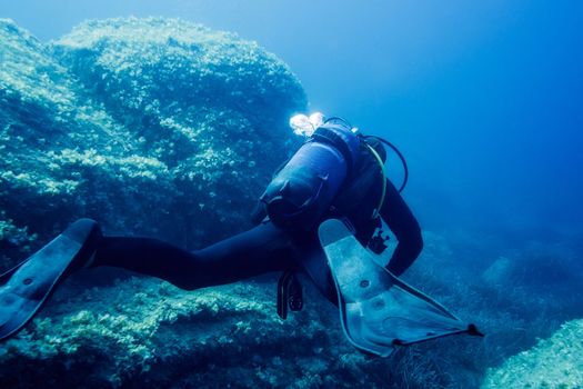 diver submerged near the bottom rocks, the water is blue and turquoise, the stones are covered with small algae. The diver turns his back to the camera and his fins are in the foreground