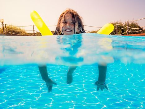 Underwater view of a girl swimming smiling toward the camera with her yellow float in the pool, behind her the glow of the summery sun gleams golden