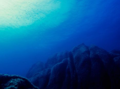 large rocks of the seabed lit up slightly by the sun reflected on the surface of the sea, small fish swim in crystal clear blue and turquoise water