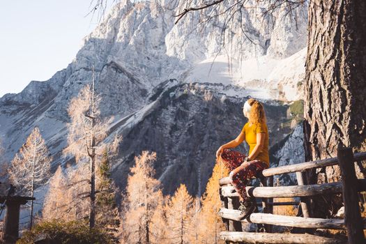 Side view of young caucasian woman hiker in colorful clothes, hiking gear, sitting on a wooden fence somewhere up in the mountains, enjoying the view of the mountain peaks on a beautiful sunny autumn day.