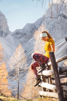 Side view of young caucasian woman hiker in colorful clothes, hiking gear, sitting on a wooden fence somewhere up in the mountains, enjoying the view of the mountain peaks on a beautiful sunny autumn day.