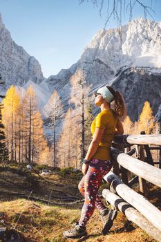 Side view of young caucasian woman hiker in colorful clothes, hiking gear, leaning on a wooden fence somewhere up in the mountains, enjoying the view of the mountain peaks on a beautiful sunny autumn day.