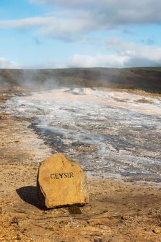 Vertical photo of steam coming up from geyser and hot water spring in Iceland. A stone with a sign geyser on it in front.