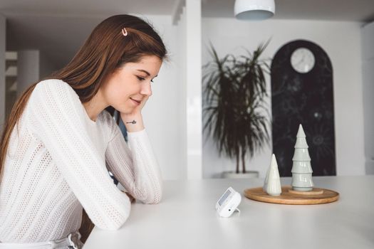 Young caucasian woman dressed in white sitting by the white kitchen island looking at the baby monitor, watching her baby sleep.