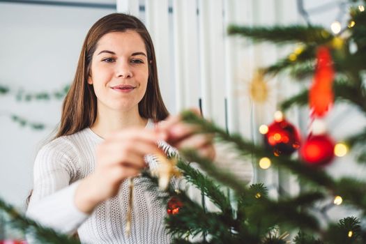 Beautiful smiling cheerful caucasian woman dressed in white outfit decorating a natural Christmas tree, putting red ornaments on the tree, holding a red Christmas ornament.