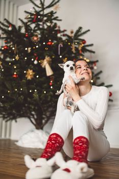 Smiling caucasian woman sitting under the Christmas tree dressed in white with a little white chihuahua in her hands. Vertical photo.