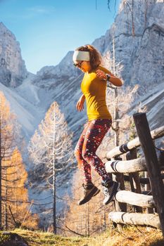 Side view of young caucasian woman hiker in colorful clothes, hiking gear, jumping of a wooden fence somewhere up in the mountains, enjoying the view of the mountain peaks on a beautiful sunny autumn day.