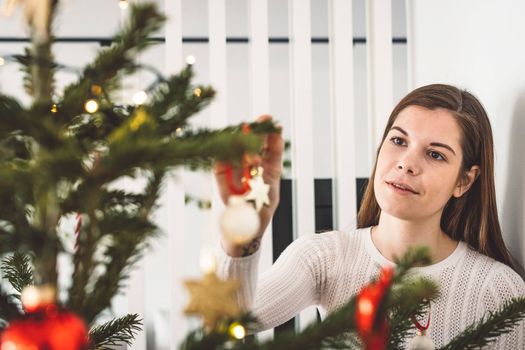 Beautiful smiling cheerful caucasian woman dressed in white outfit decorating a natural Christmas tree, putting red ornaments on the tree, holding a red Christmas ornament.