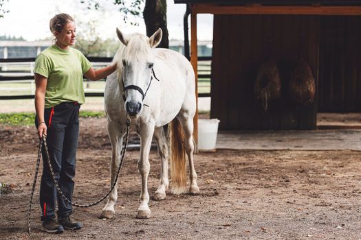 Caucasian woman trainer holding a white horse on the leash, taking hime horseback riding on the ranch.