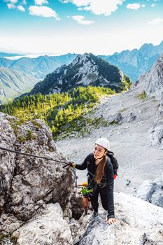Caucasian woman mountaineer climbing up the mountain on via ferrata trail, high up in the mountain peaks. Woman wearing a protective head gear and uses a safety harness while she climbs.