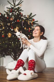 Smiling caucasian woman sitting under the Christmas tree dressed in white with a little white chihuahua in her hands. Vertical photo.