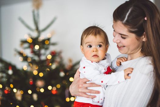 Portrait of young mom and her daughter, baby girl having fun on Christmas, decorating the Christmas tree in festive outfits. Smiling baby girl playing with her mom.