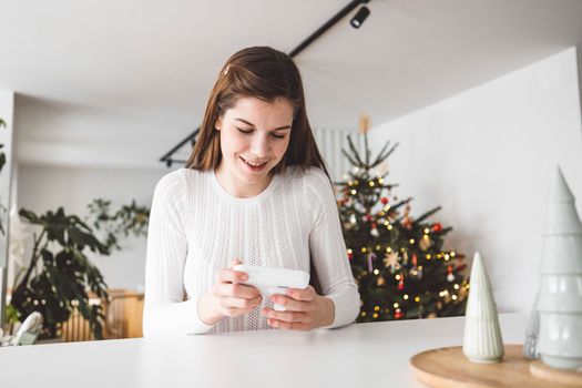 Young caucasian woman dressed in white sitting by the white kitchen island looking at the baby monitor, watching her baby sleep.