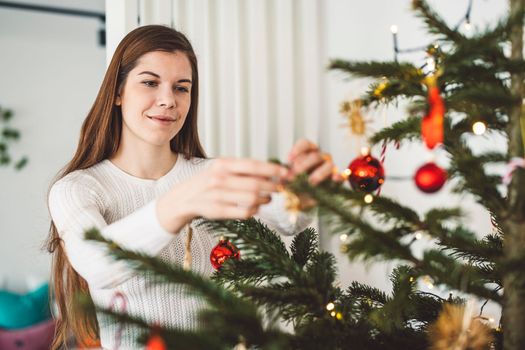 Beautiful smiling cheerful caucasian woman dressed in white outfit decorating a natural Christmas tree, putting red ornaments on the tree, holding a red Christmas ornament.