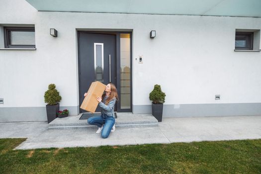 Thrilled caucasian woman sitting down at the front door with a big cardboard box that just came in the mail. Woman receiving an exciting package.