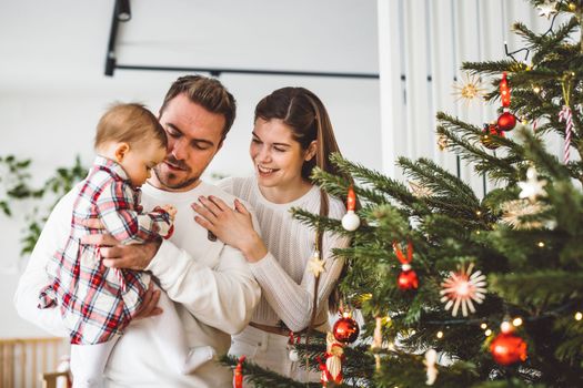 Happy young cheerful caucasian family of three mom dad and baby girl having fun decorating the Christmas tree. Family Christmas portrait.