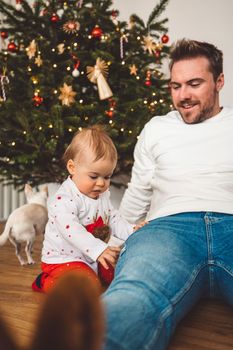 Father and daughter, baby girl having fun on Christmas, decorating the Christmass tree in festive outfits. Smiling baby girl playing with her dad.