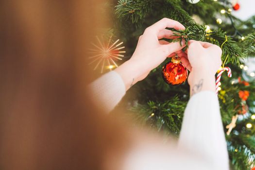 Unrecognizable caucasian woman decorating the Christmas tree, putting red ornament on the tree, holding a red Christmas ornament.