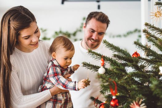 Happy young cheerful caucasian family of three mom dad and baby girl having fun decorating the Christmas tree. Family Christmas portrait.