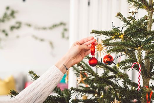 Unrecognizable caucasian woman decorating the Christmas tree, putting red ornament on the tree, holding a red Christmas ornament.