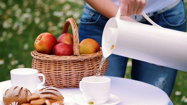 On the background of a chamomile lawn, women's hands pour tea from a white jug into a white cup, a thermos bottle. On the table there is also a basket with red apples. High quality photo
