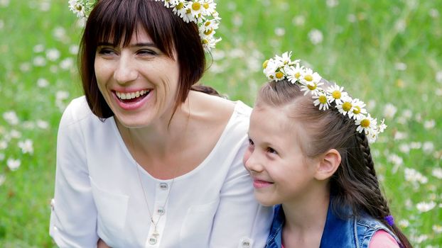 Summer, amidst a chamomile lawn, in the forest, a young woman, a brunette and a girl of seven, mother and daughter weave a wreath of chamomiles, laughing, trying on wreaths. High quality photo