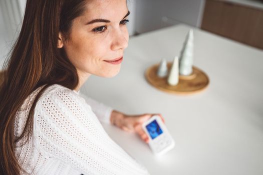 Young caucasian woman dressed in white sitting by the white kitchen island looking at the baby monitor, watching her baby sleep.