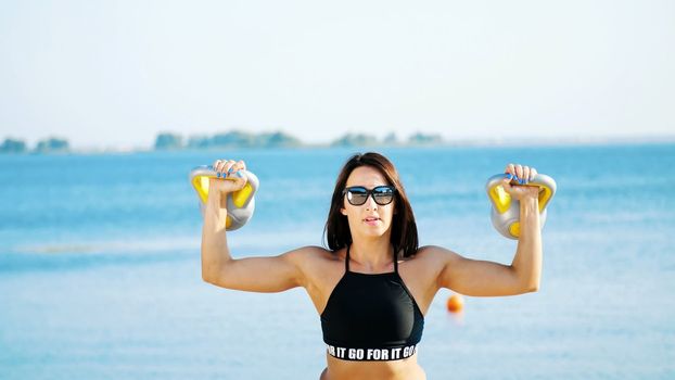 Beautiful, athletic, sexy young woman, in sunglasses, in swimsuit, coach, instructor, performs exercises with weights. On the beach, near the sea, river, in summer, in sun rays. High quality photo