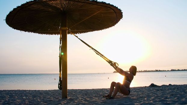 athletic, sexy young woman in swimsuit, coach, instructor, performs, doing exercises . On the beach, summer, sunset, in sun rays. High quality photo