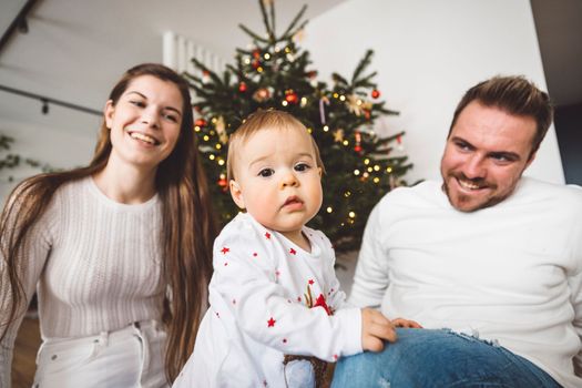 Happy young cheerful caucasian family of three mom dad and baby girl having fun decorating the Christmas tree. Family Christmas portrait. 