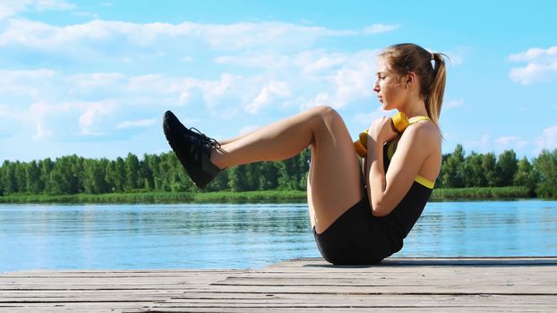Beautiful, athletic young blond woman stretching, swings press. Lake, river, blue sky and forest in the background, summer sunny day. High quality photo