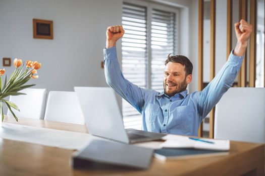 Excited caucasian man cheering for his team, watching a football game on his laptop, man celebrating a win with clenched fists. 