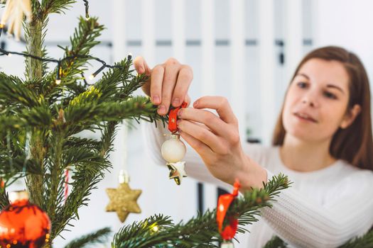 Beautiful smiling cheerful caucasian woman dressed in white outfit decorating a natural Christmas tree, putting red ornaments on the tree, holding a red Christmas ornament. 