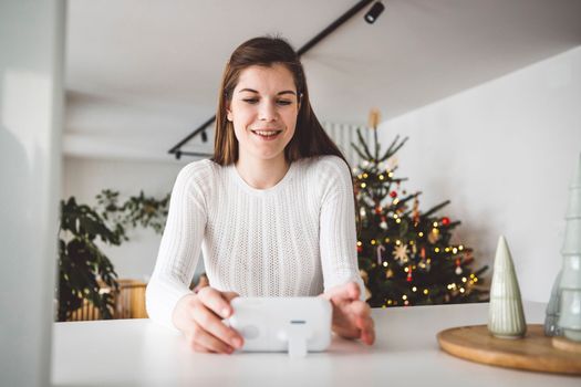 Young caucasian woman dressed in white sitting by the white kitchen island looking at the baby monitor, watching her baby sleep. 