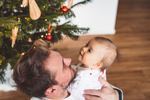 Father and daughter, baby girl having fun on Christmas, decorating the Christmass tree in festive outfits. Smiling baby girl playing with her dad. 