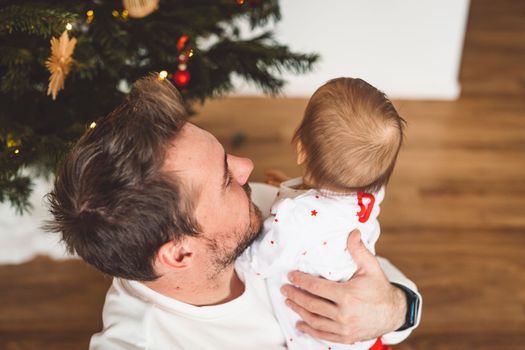 Father and daughter, baby girl having fun on Christmas, decorating the Christmass tree in festive outfits. Smiling baby girl playing with her dad. 