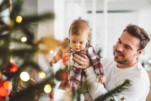 Father and daughter, baby girl having fun on Christmas, decorating the Christmass tree in festive outfits. Smiling baby girl playing with her dad. 