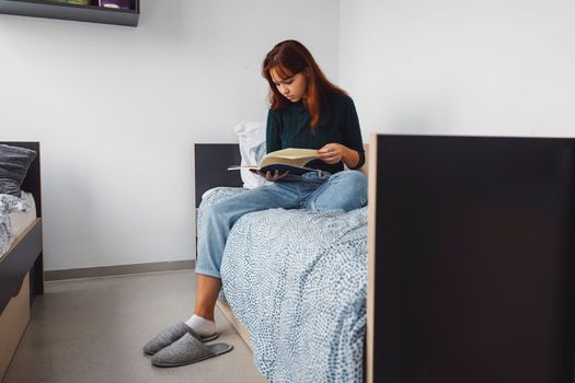 Young caucasian woman, college student studying in her dorm room, sitting on the bed. Bright room with lots of natural light. 