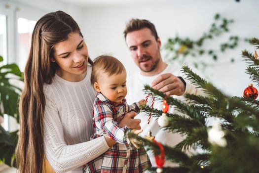 Happy young cheerful caucasian family of three mom dad and baby girl having fun decorating the Christmas tree. Family Christmas portrait. 