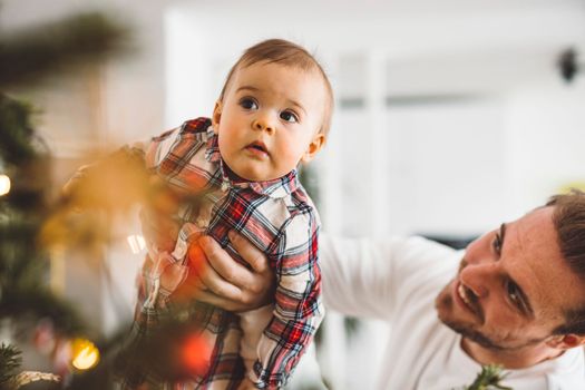 Father and daughter, baby girl having fun on Christmas, decorating the Christmass tree in festive outfits. Smiling baby girl playing with her dad. 