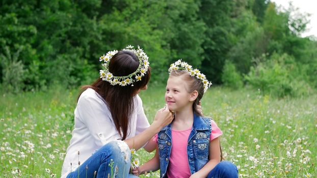 Summer, amidst a chamomile lawn, in the forest, a young woman, a brunette and a girl of seven, mother and daughter weave a wreath of chamomiles, laughing, trying on wreaths. High quality photo